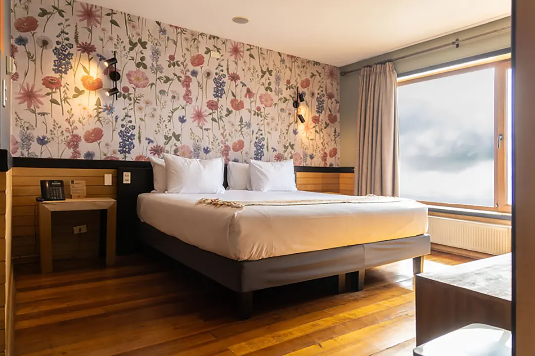 Vista de una acogedora habitación doble en un hotel pet friendly en Puerto Varas, que muestra una cama de matrimonio hecha frente a un gran ventanal con cortinas beige y un radiador debajo.