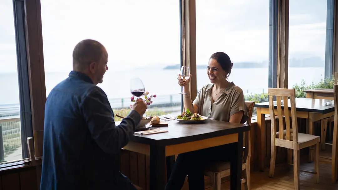 Una pareja sonriente en el restaurante de Hotel Bellavista brindando con vino frente a una gran ventana con vista al Lago Llanquihue, disfrutando de actividades en Puerto Varas.